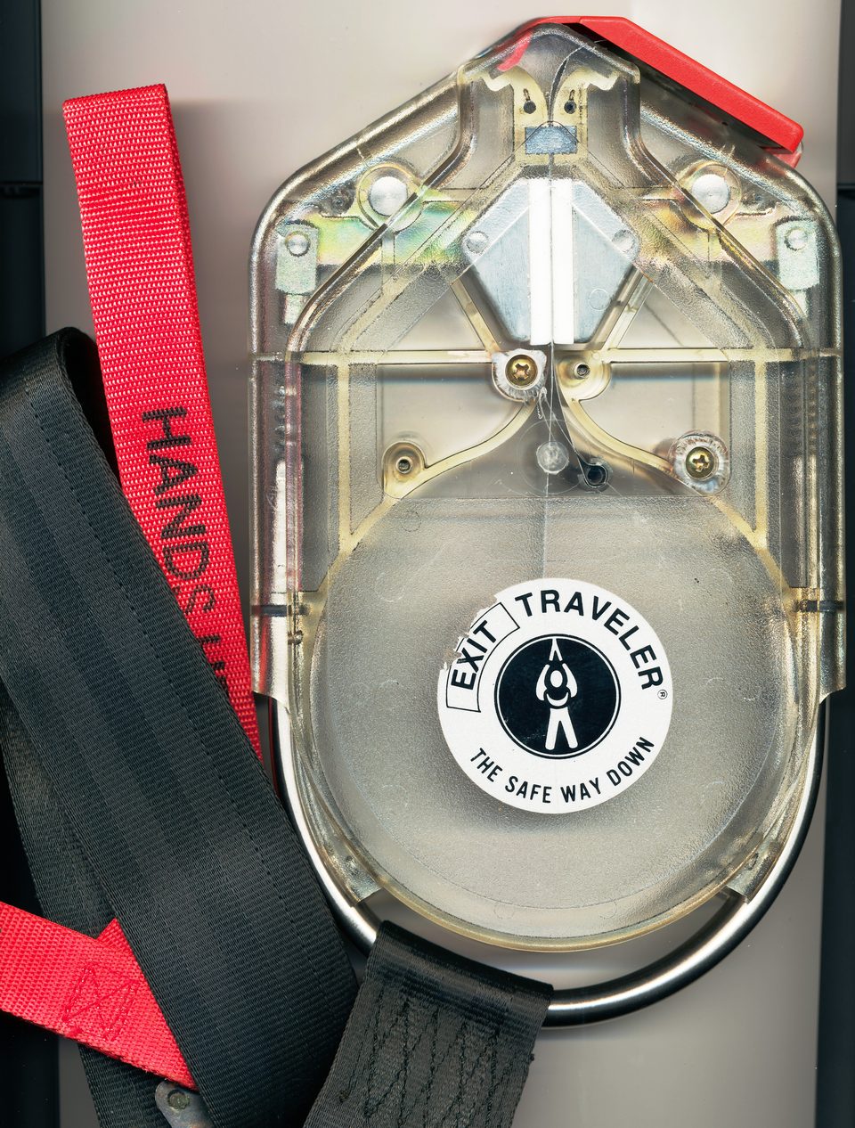 Close-up of a see-through Exit Traveler housing with a round EXIT TRAVELER label, a metal loop at the base, a red strap and black webbing