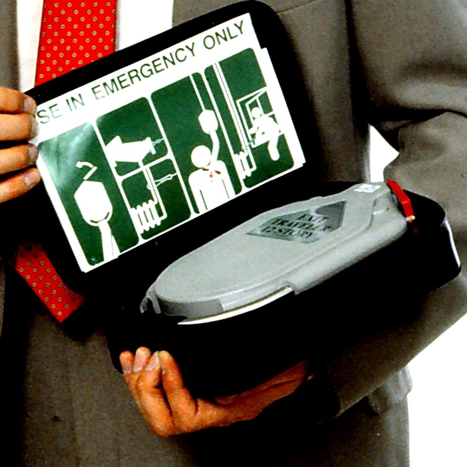 A hand holding an open black case; the lid shows a green pictorial instruction panel and the grey Exit Traveler unit sits inside