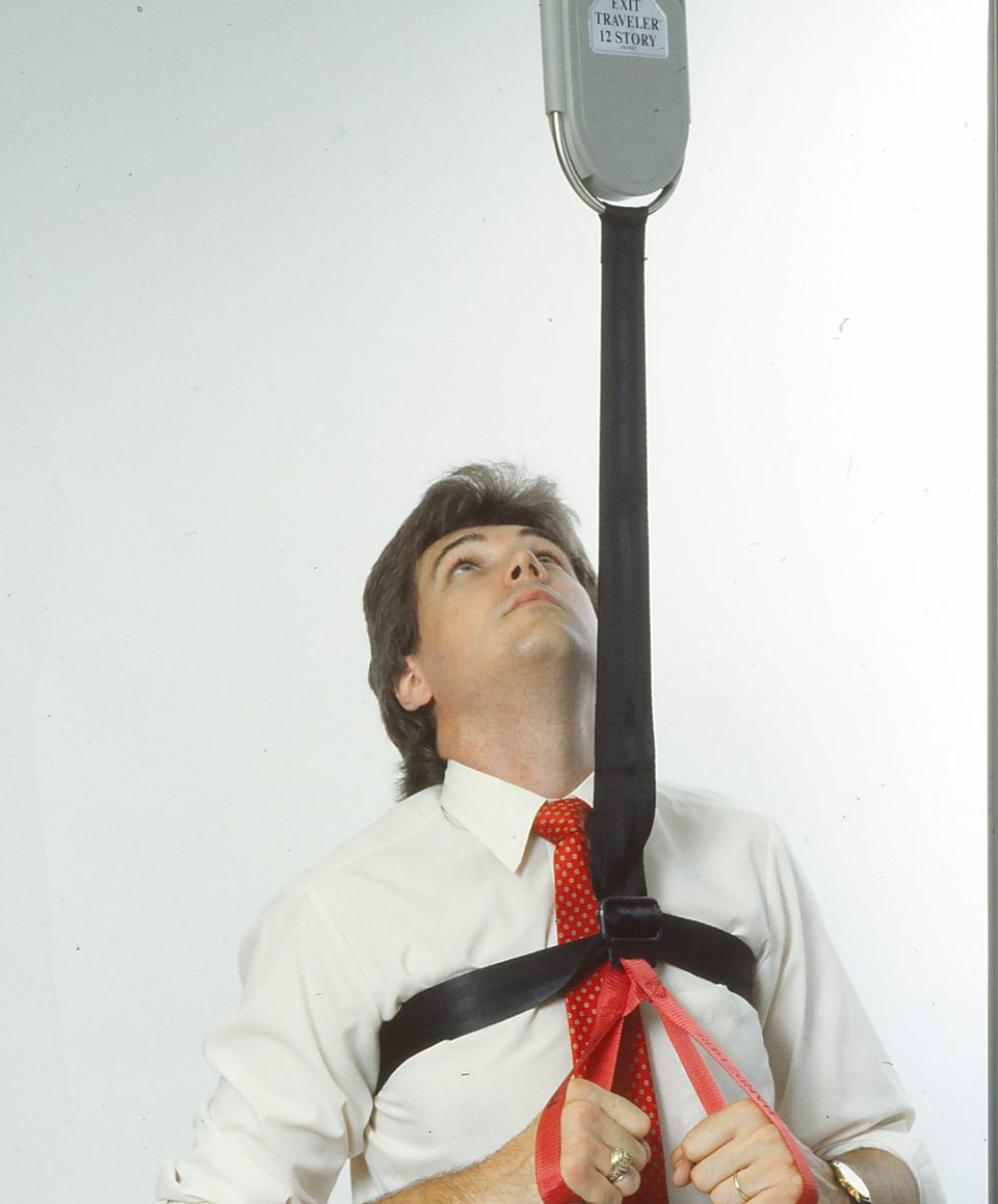 Rick Denzien in a white shirt and red tie wearing a black and red harness, looking up at the grey Exit Traveler unit hanging above him on a black strap