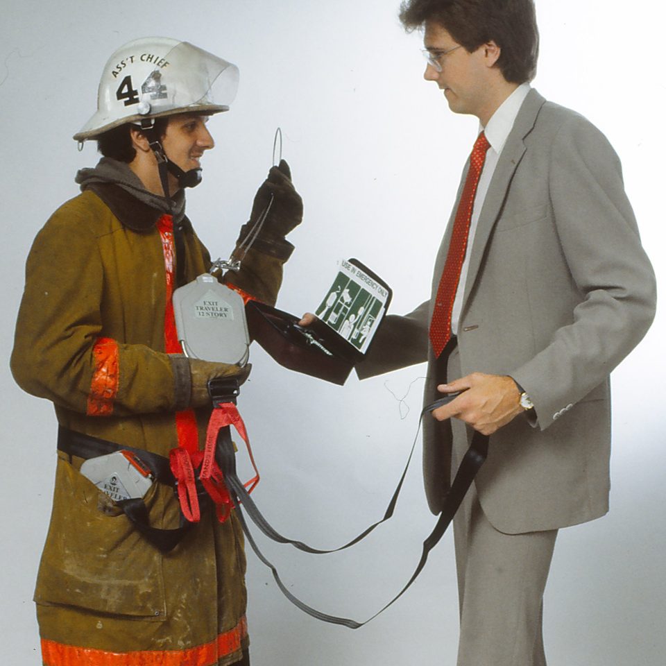 A firefighter in a white helmet and turnout coat wearing a red harness, facing a man in a grey suit who holds the Exit Traveler unit and its black cable