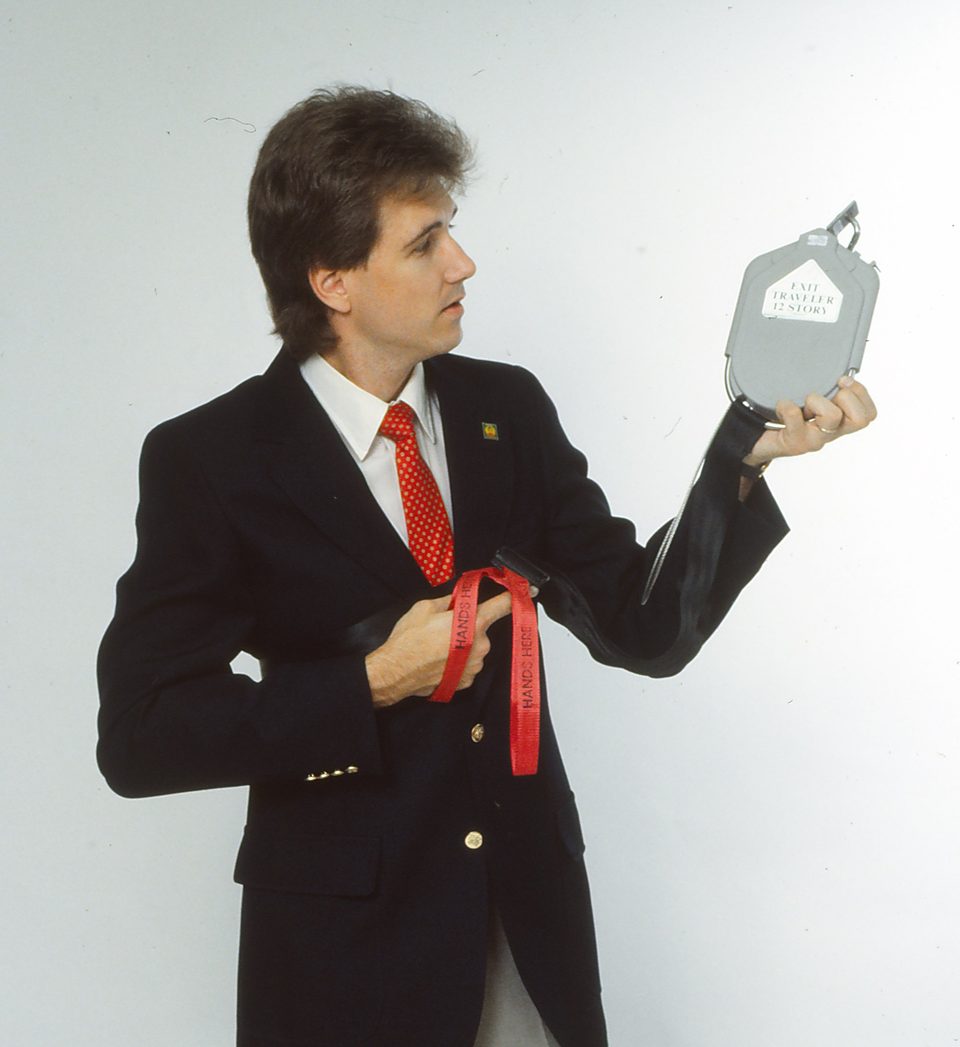 Rick Denzien in a dark jacket and red tie holding the grey Exit Traveler unit, with its red harness strap in front of him