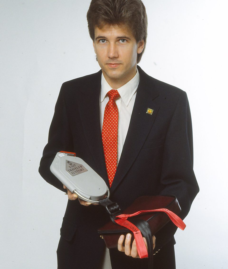 Rick Denzien in a dark blazer and red tie holding the grey Exit Traveler unit and a folded black harness with a red strap
